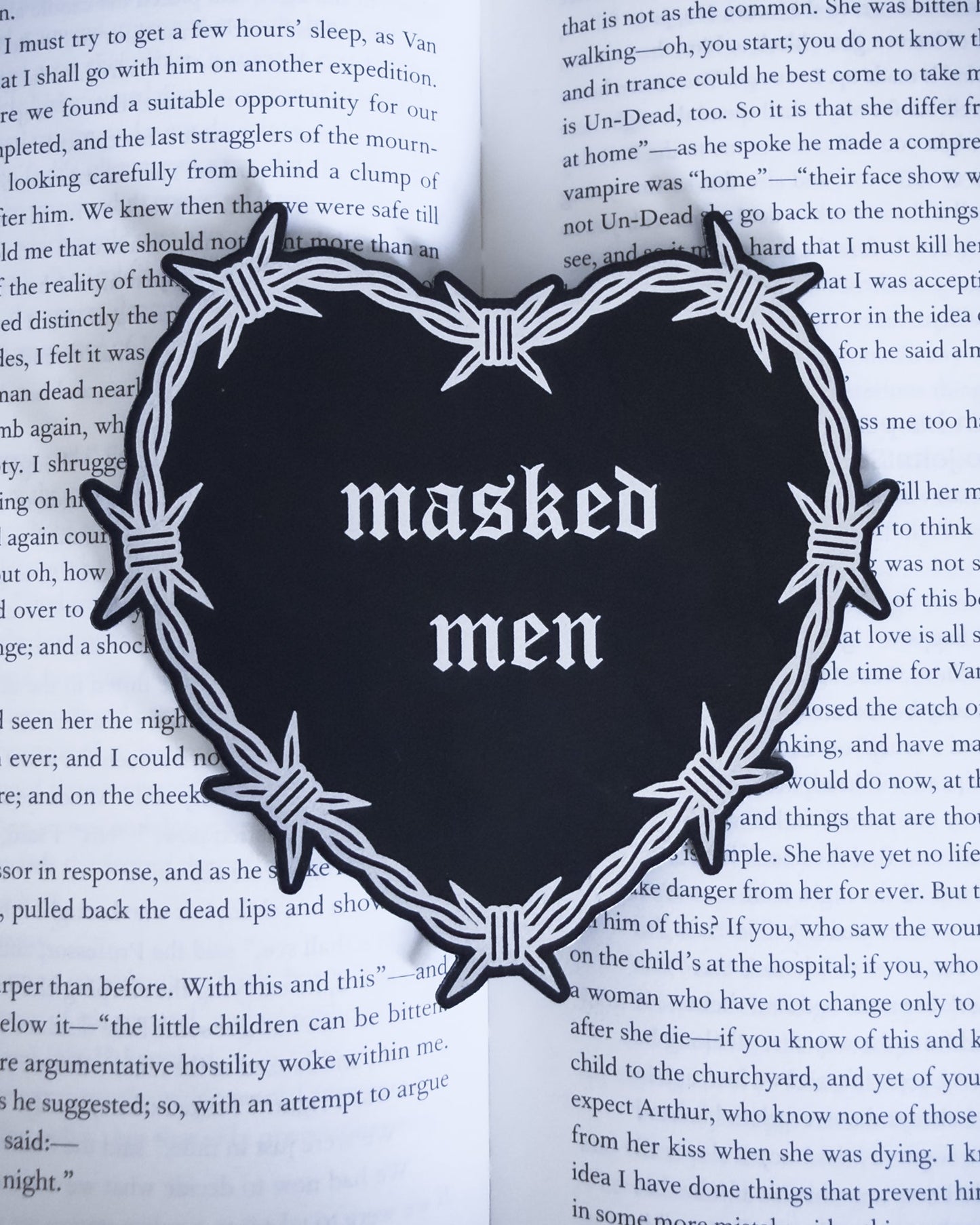 Barbed wire heart bookmark with “masked men” text in black and silver leatherette, resting on an open book