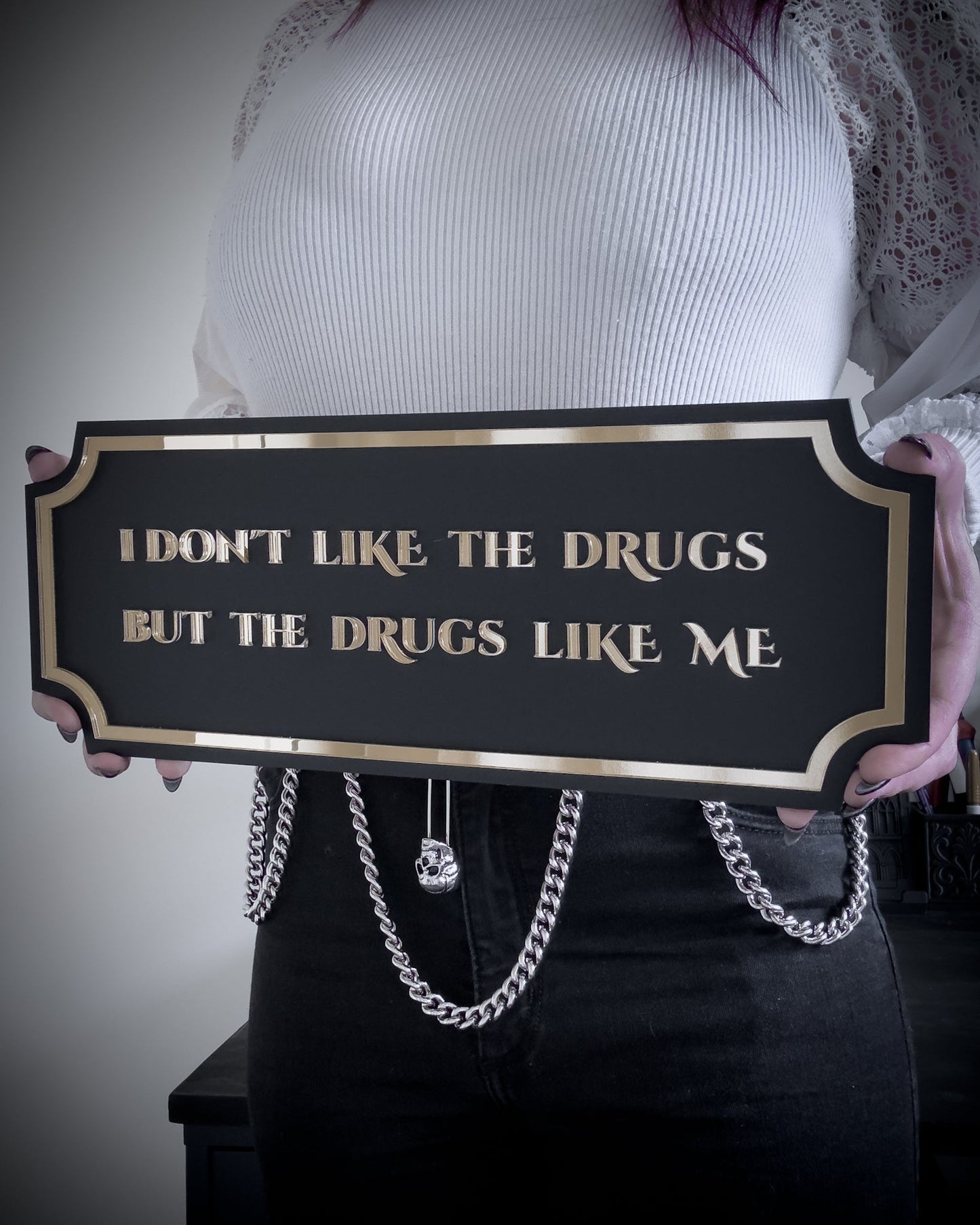 Black gothic wall plaque with gold lettering reading "I don't like the drugs but the drugs like me" held by person