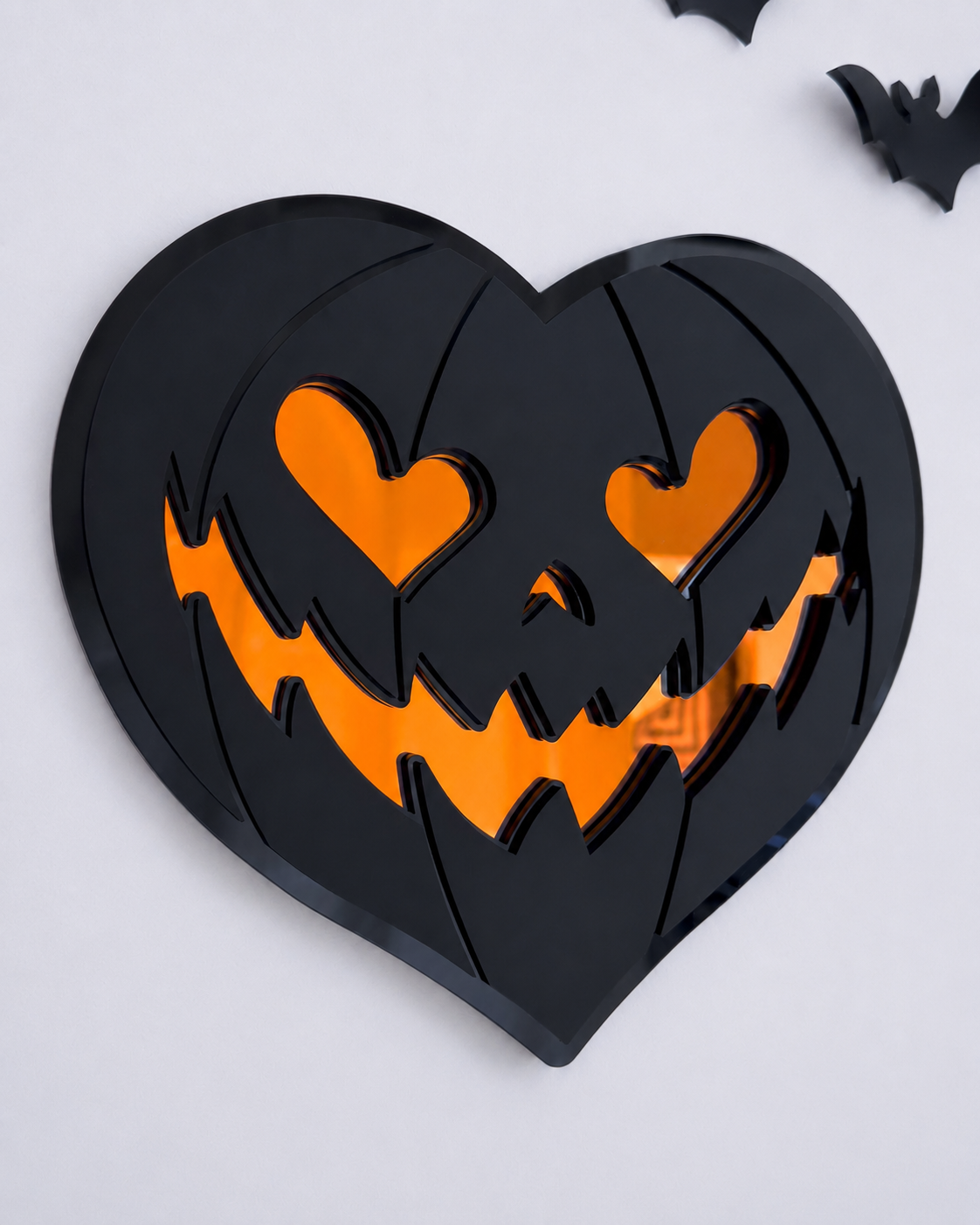 Angled view of black heart-shaped pumpkin wall art with orange mirrored eyes and smile on a bright cool-toned wall.