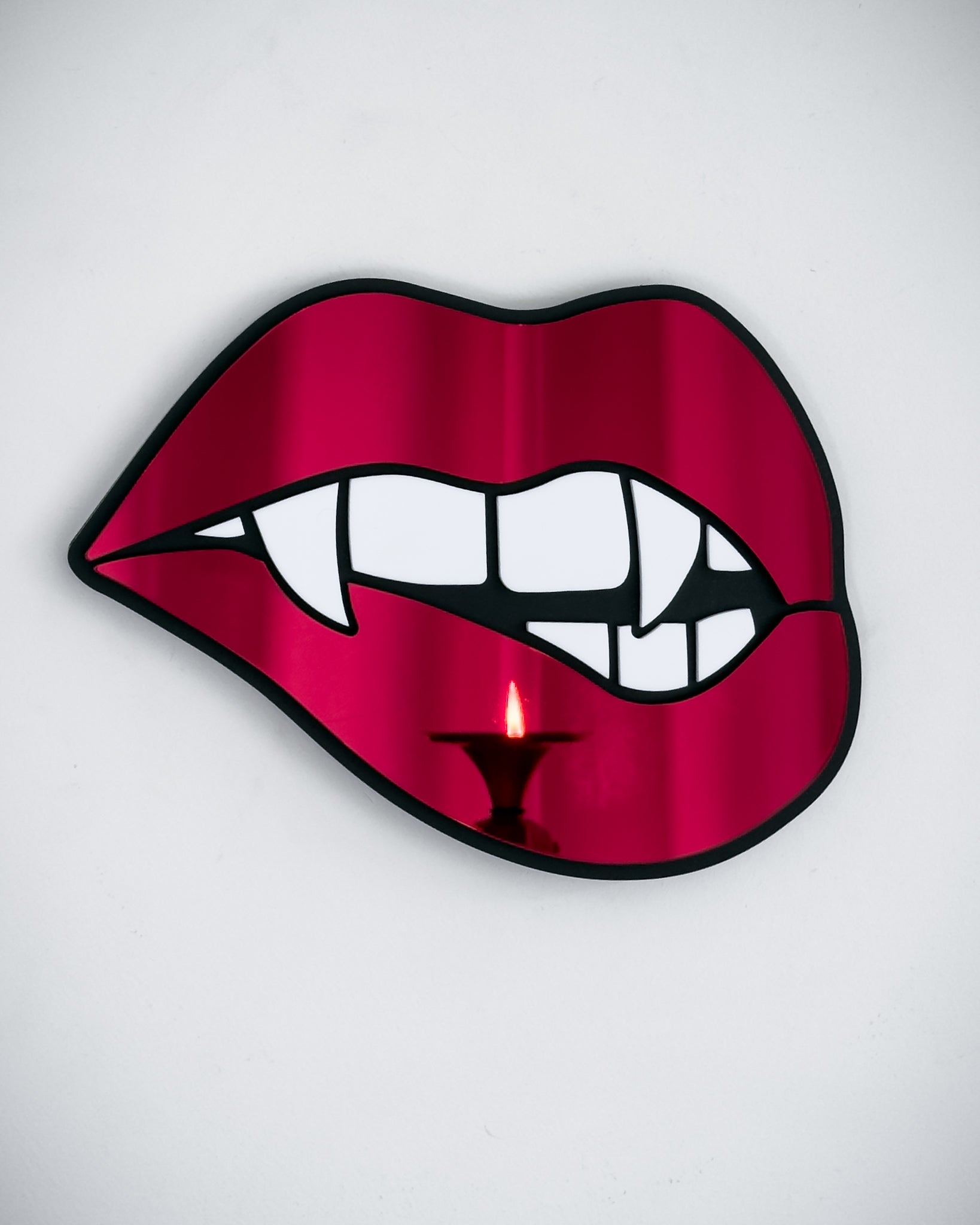 Vampire lips mirror in red acrylic with fangs, reflecting candlelight on a white wall