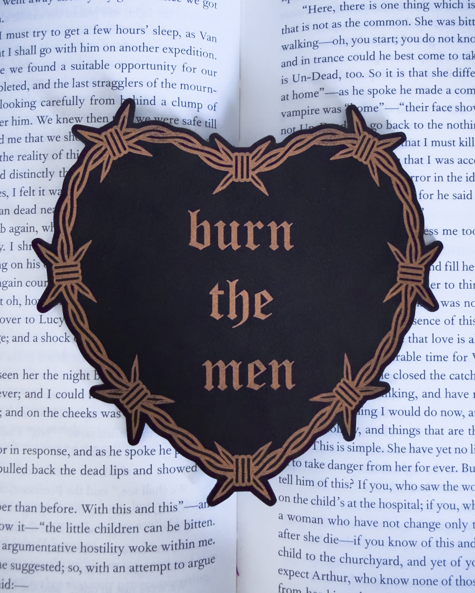 Barbed wire heart bookmark with “burn the men” text in black and copper leatherette, resting on an open book