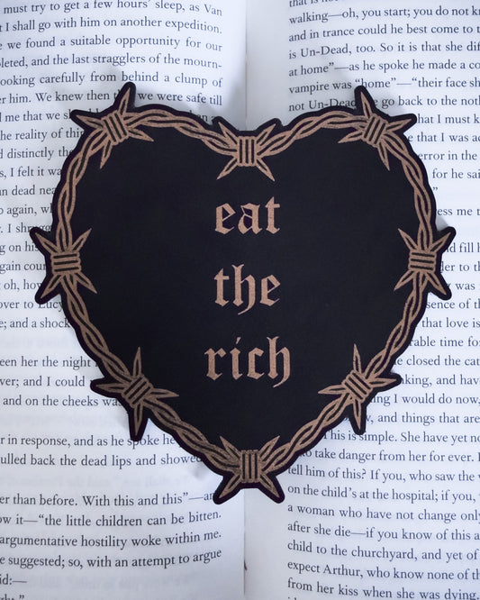 Barbed wire heart bookmark with “eat the rich” text in black and copper leatherette, resting on an open book