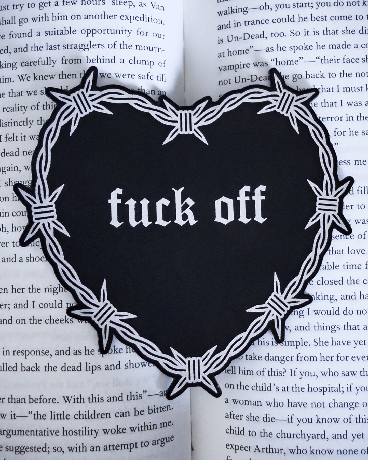 Barbed wire heart bookmark with “fuck off” text in black and silver leatherette, resting on an open book