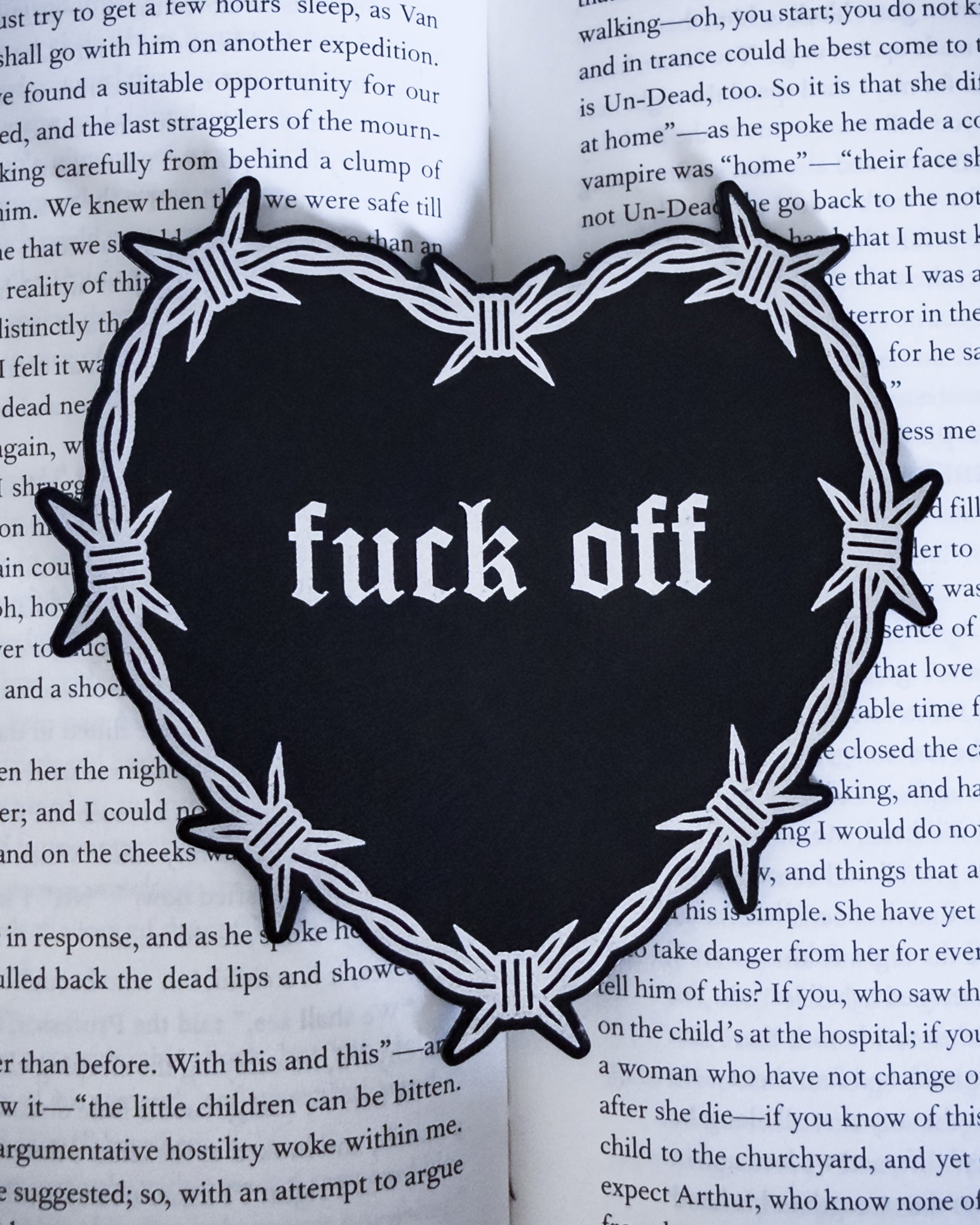 Barbed wire heart bookmark with “fuck off” text in black and silver leatherette, resting on an open book