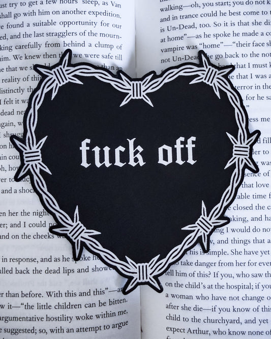 Barbed wire heart bookmark with “fuck off” text in black and silver leatherette, resting on an open book