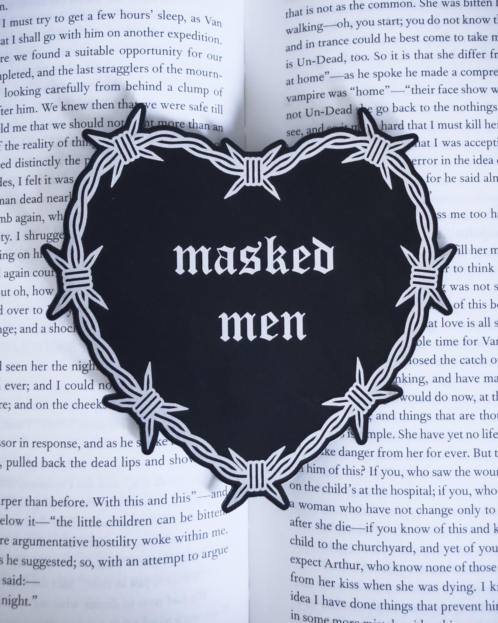 Barbed wire heart bookmark with “masked men” text in black and silver leatherette, resting on an open book