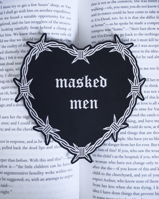 Barbed wire heart bookmark with “masked men” text in black and silver leatherette, resting on an open book