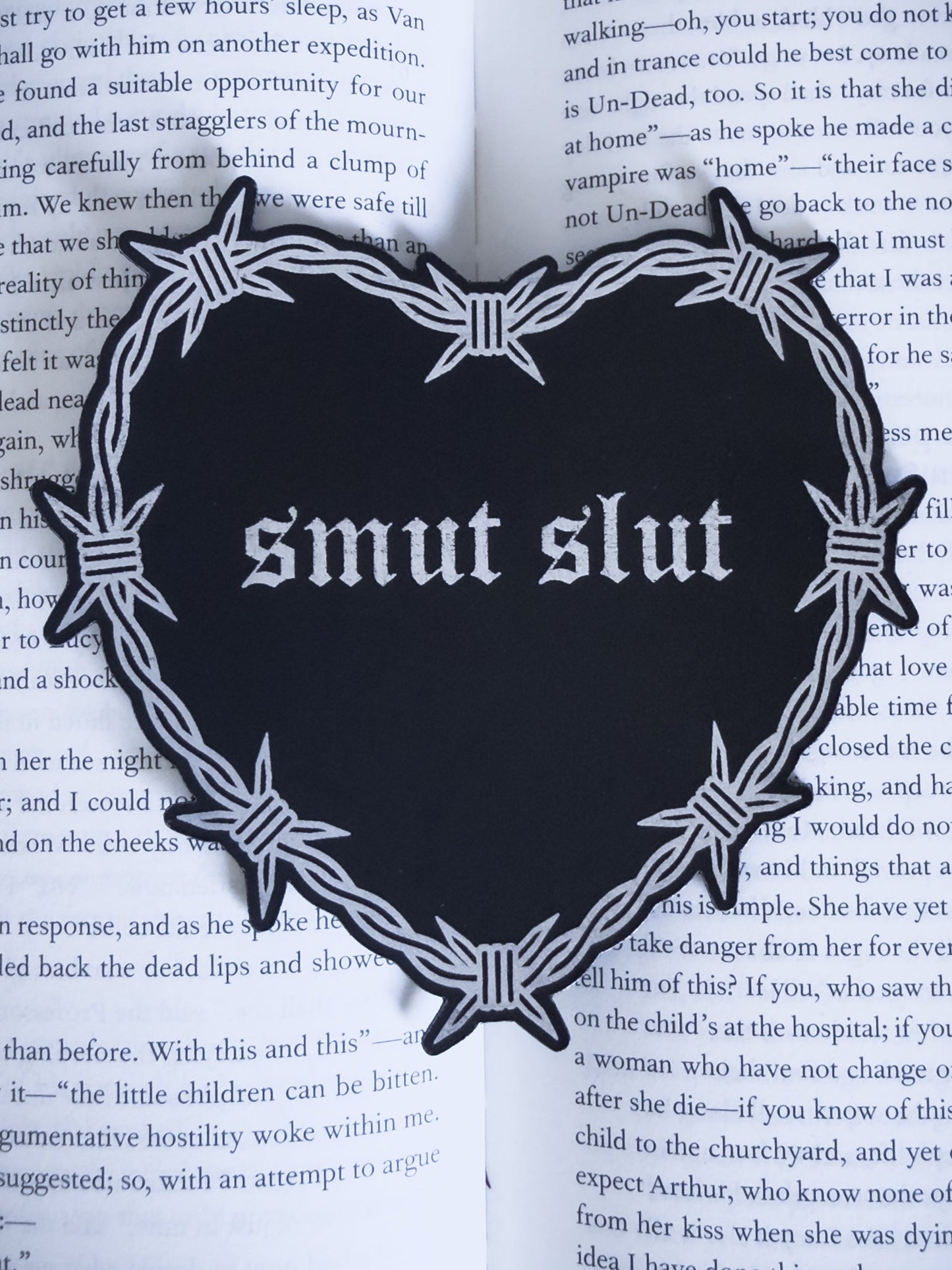Barbed wire heart bookmark with “smut slut” text in black and silver leatherette, resting on an open book