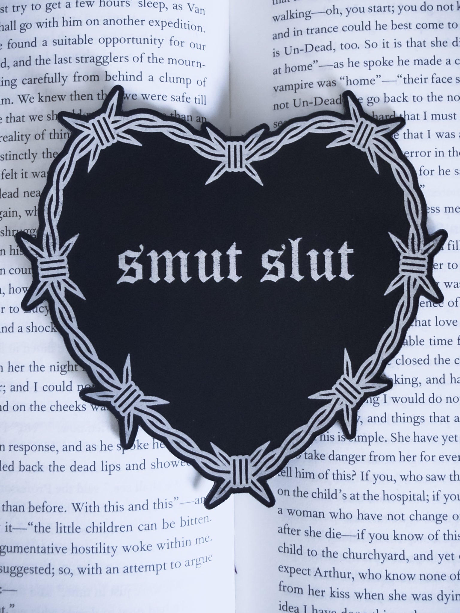 Barbed wire heart bookmark with “smut slut” text in black and silver leatherette, resting on an open book