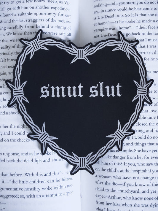 Barbed wire heart bookmark with “smut slut” text in black and silver leatherette, resting on an open book