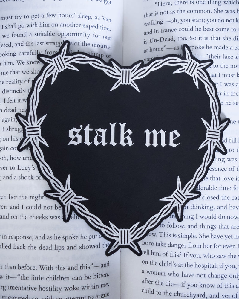 Barbed wire heart bookmark with “stalk me” text in black and silver leatherette, resting on an open book