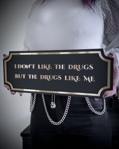 Black gothic wall plaque with gold lettering reading "I don't like the drugs but the drugs like me" held by person