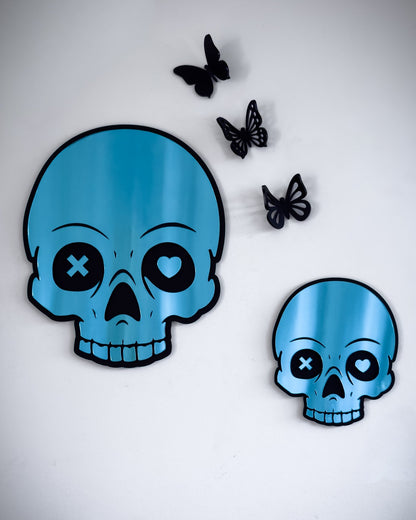 Small and large blue skull mirrors with mismatched heart and X eyes, styled with butterfly accents on a white wall