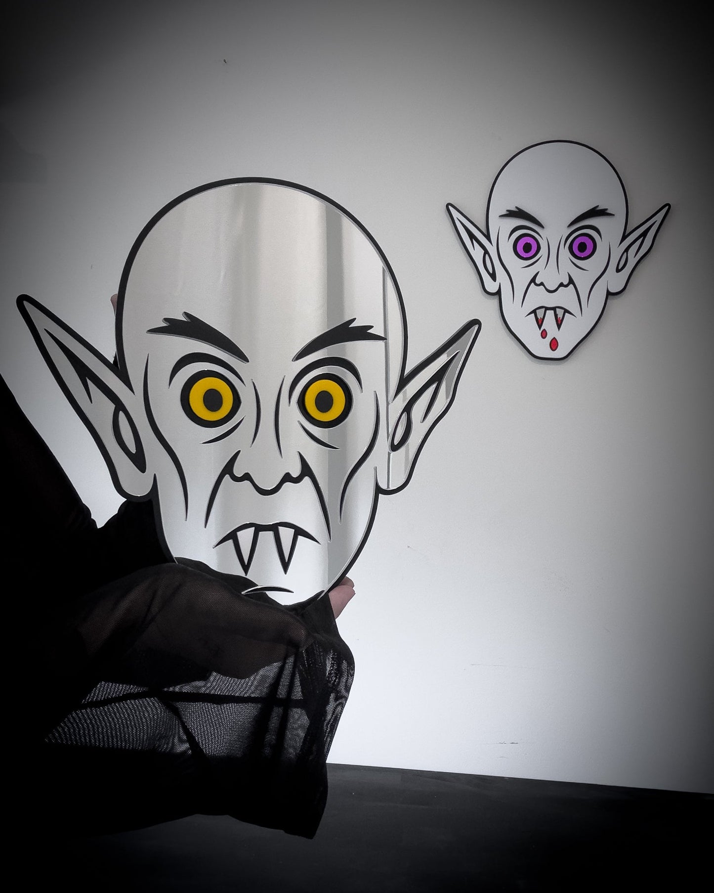 Large Gothic Nosferatu mirror with yellow eyes held for size reference, with white Nosferatu wall art on the wall behind